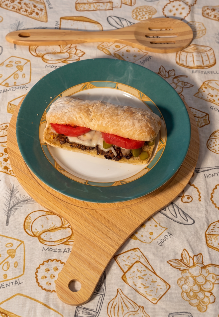 Homemade Italian ground beef sandwich with melted cheddar and gouda cheese, fresh tomatoes, pickles, ketchup, and mustard on toasted Italian bread.